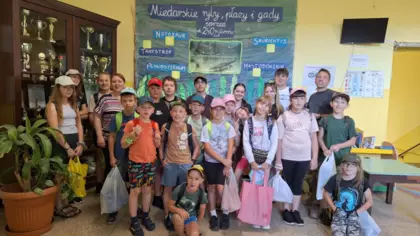 Dzieci z opiekunami stoją w szkolnym korytarzu na tle wystawy paleontologicznej o skamieniałościach sprzed 240 mln lat temu.