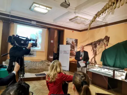 Zdjęcie z otwarcia odnowionej wystawy skamieniałości z Lisowic. Kamerzysta kręcący wypowiedź Dyrektor Instytutu Paleobiologii w Warszawie. W tle szkielet dinozaura.