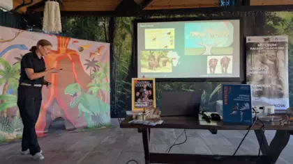 Na zdjęciu stoi kobieta w czarnym ubraniu. Trzyma wskaźnik laserowy. Za nią widać fotościankę z wybuchającym wulkanem, palmami i dinozaurami. Na ścianie jest ekran wyświetlaną prezentacją o słoniu leśnym. Na pierwszym planie widać drewnianą ławę okładkami czasopism naukowych. Po prawej stronie, z tyłu stoi roll-up z reklamą Muzeum Paleontologicznego w Lisowicach.