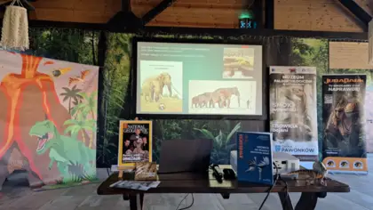 Na zdjęciu widać fotościankę z wybuchającym wulkanem, palmami i dinozaurami. Na ścianie jest ekran wyświetlaną prezentacją o słoniu leśnym. Na pierwszym planie widać drewnianą ławę okładkami czasopism naukowych. Po prawej stronie, z tyłu stoi roll-up z reklamą Muzeum Paleontologicznego w Lisowicach.