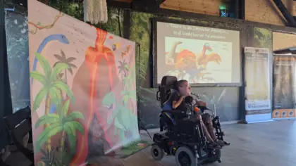 Na zdjęciu widać młodego mężczyznę na wózku inwalidzkim. Za nim stoi fotościanka z wybuchającym wulkanem, palmami i dinozaurami. Na ścianie jest duży ekran z wyświetlaną prezentacją o dinozaurach.