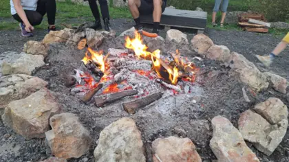 Na zdjęciu widać ognisko otoczone kamiennym kręgiem. Nad ogniskiem nachylają się ludzie pieczący kiełbaski i kromki chleba nad ogniskiem.