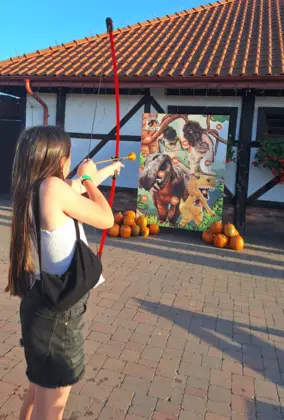 Na zdjęciu widać dziewczynkę o długich, ciemnych włosach, która strzela z łuku do planszy z dinozaurami.