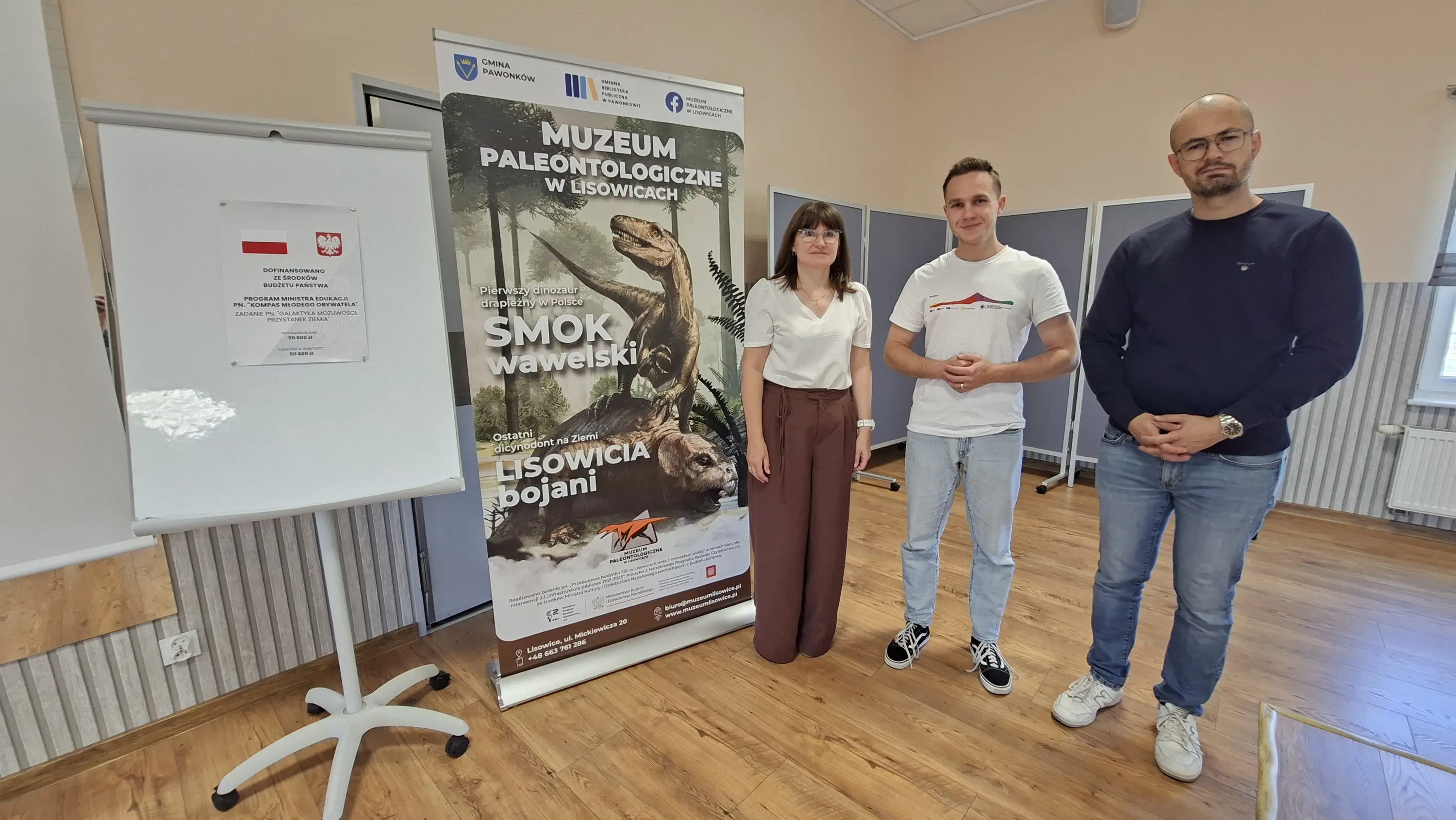Dyrektor Gminnej Biblioteki w Pawonkowie oraz dwaj geolodzy stoją obok roll-upu z wizerunkiem smoka i lisowicji oraz napisem „Muzeum Paleontologiczne w Lisowicach”, a obok widoczna jest tablica z plakatem projektu „Galaktyka możliwości: przystanek Ziemia”.