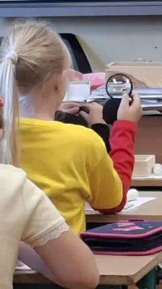 Dziewczynka w żółtej bluzce, widziana od tyłu, siedzi w ławce podczas warsztatów geologicznych. W dłoni trzyma lupę