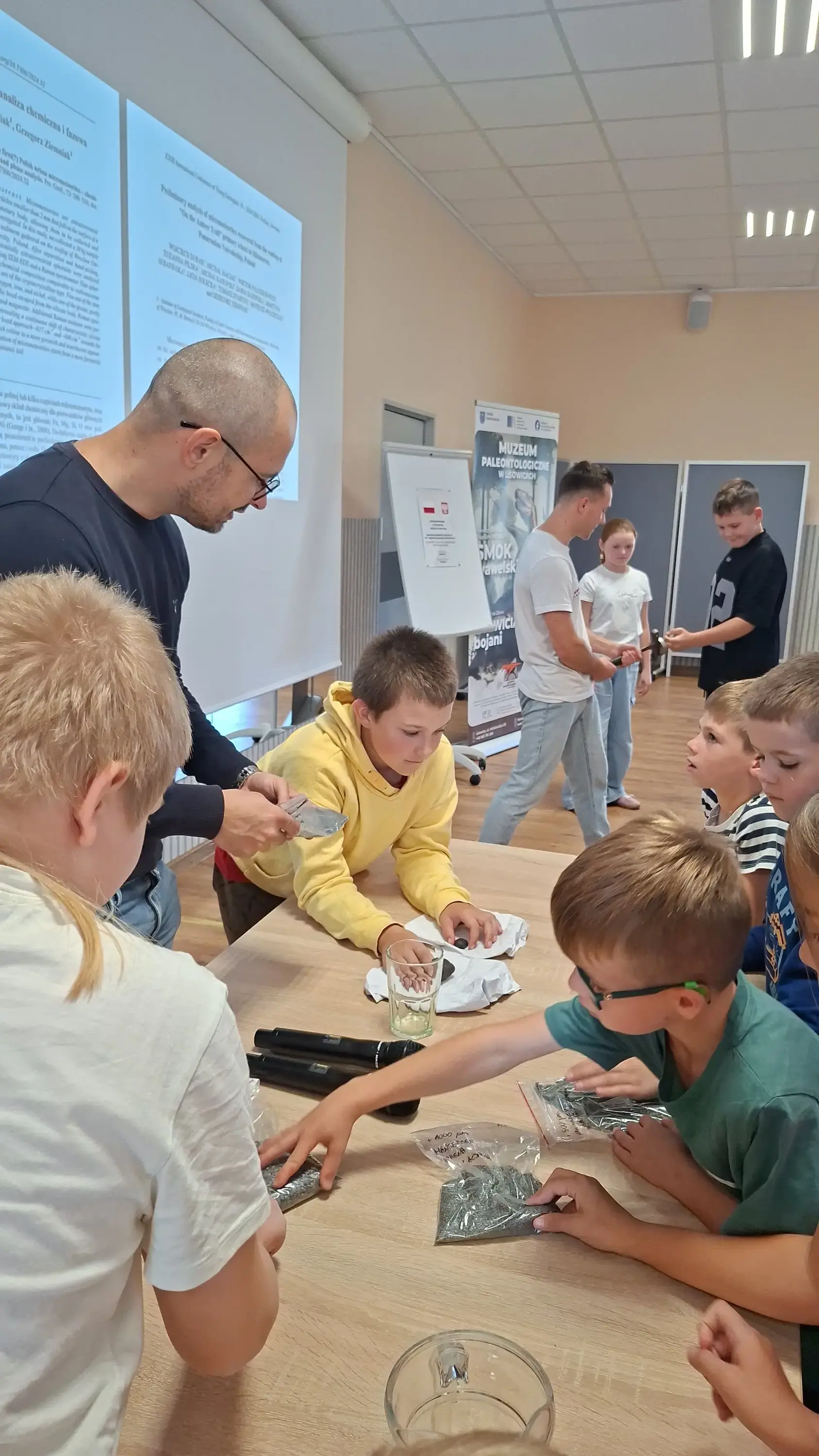 Dzieci badają próbki geologiczne przy stole pod okiem naukowca, podczas gdy w tle drugi naukowiec prezentuje działanie silnego magnesu z młotkiem, a za nimi widoczny jest ekran z prezentacją o kosmosie.