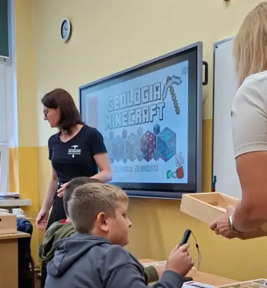 Dwóch chłopców siedzi w ławce podczas warsztatów „Geologia Minecraft”. W tle prowadząca zajęcia oraz nauczycielka trzymająca skrzynkę z materiałami, a na tablicy wyświetlony jest temat warsztatów.