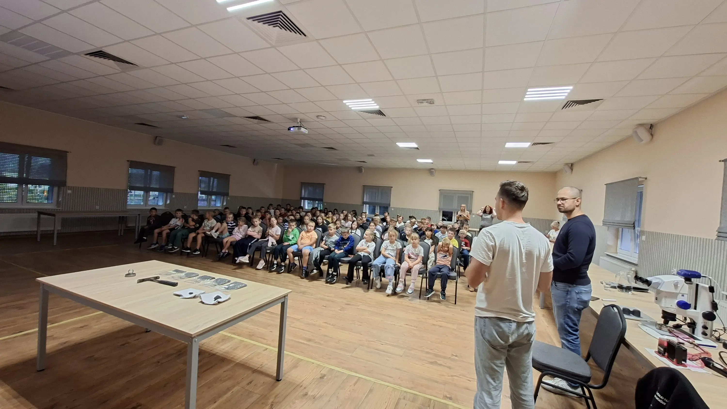 Dwóch naukowców prowadzi pokaz dla około stu dzieci z szkoły podstawowej zgromadzonych w dużej sali. Po lewej stronie na stole widoczne są próbki i młotek, a po prawej profesjonalny mikroskop. Dzieci siedzą w rzędach na krzesłach, słuchając prelegentów.