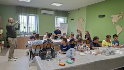 Dzieci przyglądają się skamieniałościom przy dwóch dużych stołach w sali, podczas gdy stojący po lewej stronie geolog wskazuje jedną z nich i o niej opowiada.