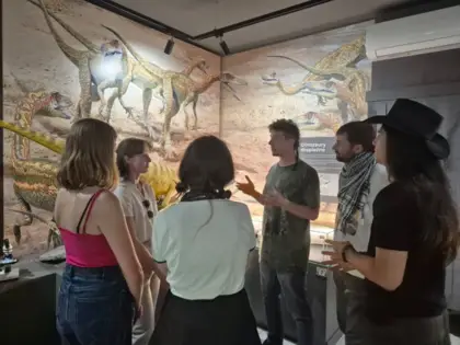 Grupka studentów stoi wokół mężczyzny, który opowiada im o wystawie. Za nimi na ścianie widać fototapetę ze stadem celofyzów.