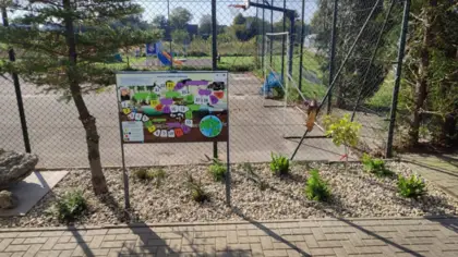 Przed ogrodzeniem z siatki metalowej wśród zielonych roślin stoi tablica z grą planszową. Za siatką widać boisko do gry w koszykówkę oraz plac zabaw.